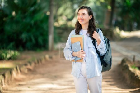 Contented european young woman student holding books and a laptop, with a gentle smile, strolling in a tranquil park setting, exuding ease and academic readiness, outdoor