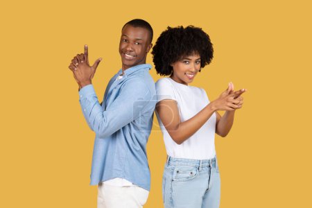 Ein afroamerikanisches Paar aus der Jahrtausendwende zeigt selbstbewusst Handzeichen mit verspieltem Gesichtsausdruck, in legerer Kleidung vor einheitlichem gelben Hintergrund, Studio