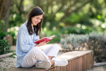 Photo for Serene european young woman student enjoying reading a red book, sitting cross-legged on a park bench, engrossed in literature with a smile in a tranquil outdoor setting, outdoor - Royalty Free Image