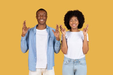 Téléchargez les photos : Un jeune couple afro-américain joyeux portant des vêtements décontractés croisent les doigts les yeux fermés sur un fond jaune, espérant bonne fortune, studio. Hope, gagne. - en image libre de droit