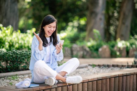 Foto de Emocionada joven estudiante europea riendo y haciendo gestos mientras mira su teléfono inteligente, sentada con las piernas cruzadas en un banco del parque de madera, disfrutando de una charla animada, al aire libre - Imagen libre de derechos