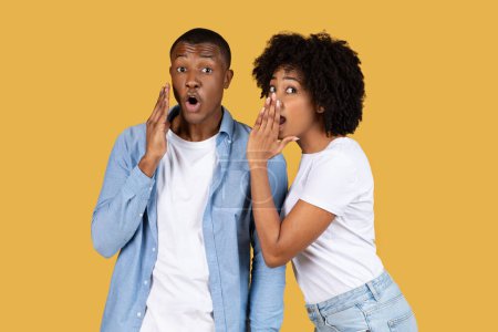 Photo for Shocked millennial African American man and woman sharing a secret, with hands to their mouths and wide-eyed expressions of surprise and gossip on a uniform yellow background - Royalty Free Image