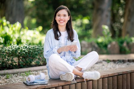 Happy european young woman student in casual wear sitting cross-legged on a park bench while holding a smartphone, with headphones and notebooks beside her, in a lush green setting
