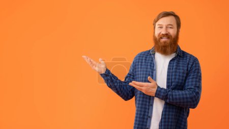Photo for Smiling Man With Red Hair And Beard Gesturing Showing Copy Space With Both Hands, Standing In Casual Plaid Shirt, Looking At Camera Over Orange Studio Background, Panorama - Royalty Free Image