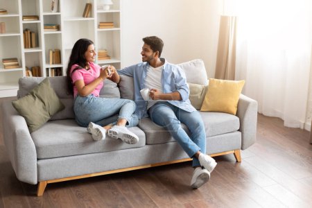 Photo for Relaxed young indian couple having pleasant conversation over coffee, comfortably sitting on sofa in well-lit living room with homely vibe, full length, free space - Royalty Free Image