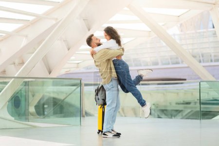 Foto de Pareja cariñosa abrazándose alegremente en la terminal del aeropuerto, hombre romántico levantando a la mujer del suelo, cónyuges alegres reunidos después de mucho tiempo separados, felices de verse, espacio de copia - Imagen libre de derechos