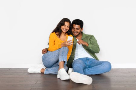 Photo for Happy Indian couple in casual clothes sitting on the floor, looking at smartphone together with joyful expressions on clean white wall background - Royalty Free Image