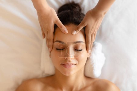Weekend at spa. Closeup of young woman receiving relaxing head massage, lying with masseuses hands on forehead indoor. Concept of relaxation therapy at luxury beauty salon