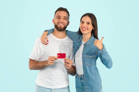 Téléchargez les photos : Joyeux jeune couple souriant et tenant la carte de crédit, approuvant l'offre bancaire avec le pouce vers le haut geste, prêt à effectuer le paiement à l'achat, embrassant sur fond bleu studio - en image libre de droit
