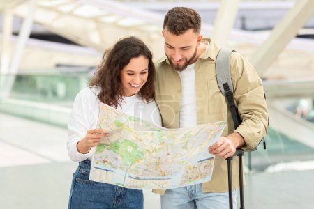 Photo for Content young couple reading city map together while standing in airport terminal, happy millennial man and woman planning vacation travel, checking journey route and smiling, free space - Royalty Free Image