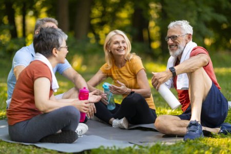 Foto de Los entusiastas del yoga jubilosos disfrutan de una charla amistosa mientras están sentados en esteras al aire libre, ancianos felices sosteniendo botellas de agua, hombres y mujeres mayores relajándose después de una sesión refrescante en el parque de verano - Imagen libre de derechos