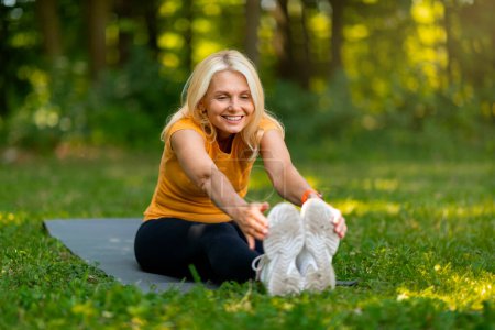 Téléchargez les photos : Femme âgée rayonnante aux cheveux blonds s'étirant vers ses orteils sur un tapis de yoga dans un parc ensoleillé, entraînement féminin âgé sportif heureux à l'extérieur, exsudant santé et bonheur, espace de copie - en image libre de droit