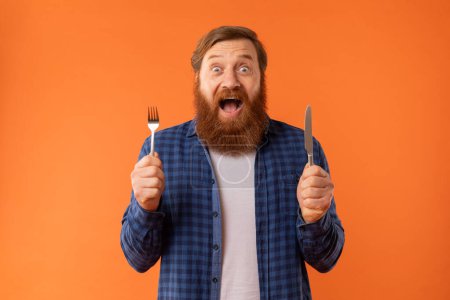 Photo for Hunger cravings. Hungry redhaired bearded man shouting holds knife and fork, expressing excitement and anticipation of delicious meal at restaurant, over orange studio backdrop, looking at camera - Royalty Free Image