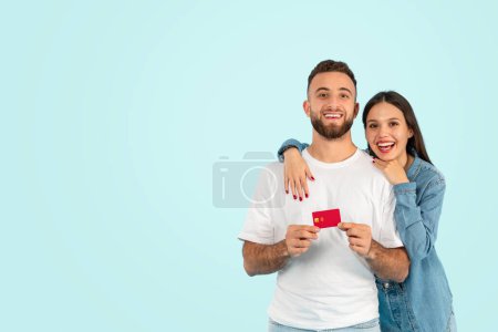 Photo for Happy European couple holds credit card, showing excitement for retail sale or bank offer against blue backdrop, embracing standing near free space for text, smiling to camera - Royalty Free Image