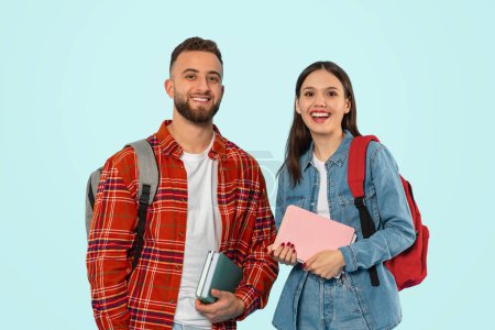 Photo for Modern students. Cheerful young couple poses casually with backpacks, textbooks and gadgets over blue wall in studio, smiling to camera while representing modern college life. Concept of education - Royalty Free Image