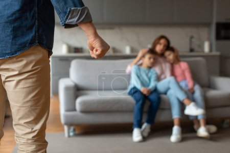 Häusliche Gewalt. Unbekannter Mann bedroht Frau und Kinder mit der Faust, verängstigte Mutter umarmt Sohn und Tochter, während sie zusammen auf Couch sitzen, selektiver Fokus auf männliche Hand