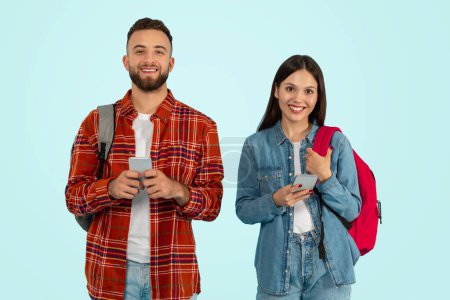 Photo for Boyfriend and girlfriend college students texting and chatting online, using cell phones while standing with backpacks over blue background, studio shot. Concept of digital communication - Royalty Free Image