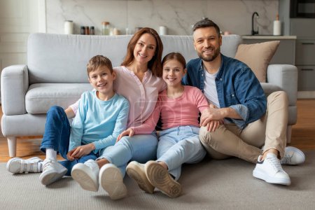 Photo for Smiling young family with preteen kids sitting on floor in cozy living room, happy parents and two children posing in comfortable home, sharing joyful and loving moment, looking at camera - Royalty Free Image
