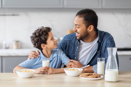 Téléchargez les photos : Heureux père afro-américain et fils préadolescent prendre le petit déjeuner ensemble, boire du lait, manger des céréales et la cuisine. Papa étreignant son enfant, avoir une conversation tout en mangeant dans la cuisine à la maison - en image libre de droit