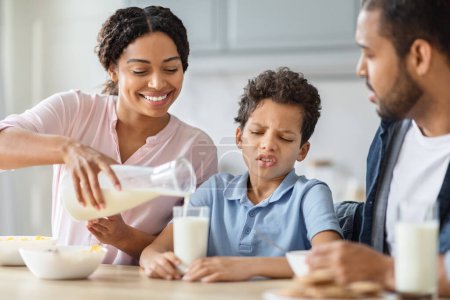 Téléchargez les photos : Un garçon afro-américain qui refuse de boire du lait. Aimer les parents noirs père mère verser des produits laitiers frais plein de calcium dans le verre de leur fils enfant, la famille ont un petit déjeuner sain ensemble - en image libre de droit