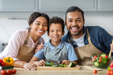 Téléchargez les photos : Portrait d'une famille afro-américaine joyeuse et aimante qui cuisine ensemble à la maison. Mère souriante, père et enfant garçon préadolescent fils préparant un repas sain à la cuisine moderne confortable, amusez-vous le week-end - en image libre de droit
