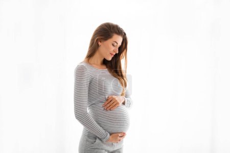 Eine junge schwangere Frau streichelt ihren Bauch, während sie vor weißem Hintergrund steht