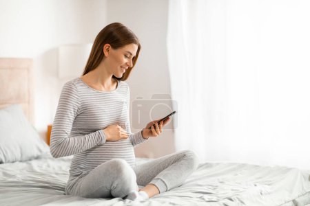 A peaceful pregnant woman engages with her phone in the comfort of her bedroom, embodying a sense of calm