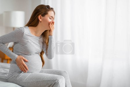 A side view of a pregnant woman holding her belly as she sits uncomfortably on a bed, depicting prenatal distress and discomfort in a home setting