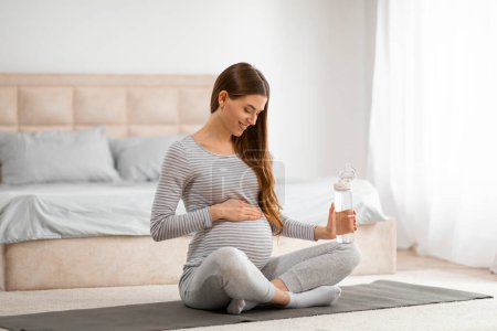 Eine ruhige werdende Mutter in einem gemütlichen Raum schlürft Wasser, während sie auf einer Yogamatte sitzt und die Flüssigkeitszufuhr hervorhebt