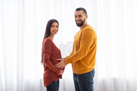 A joyful couple touches the womans pregnant belly, smiling towards camera