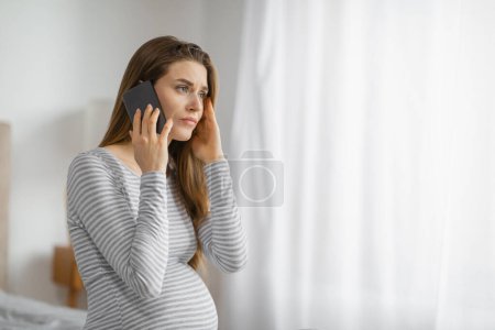 Expectant mother on phone with a worried expression, standing by a window