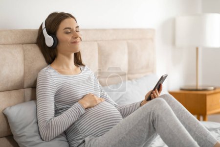 Téléchargez les photos : Confortable et contente, une femme enceinte aime la musique et navigue sur son smartphone tout en se reposant au lit - en image libre de droit