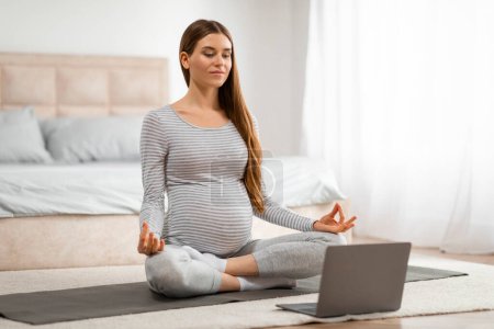 Concentrated pregnant woman seated on a yoga mat, using a laptop for online prenatal classes or relaxation