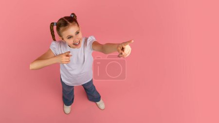 A cheerful young girl with braided hair points at copy space on a pink background, expressing fun and joy