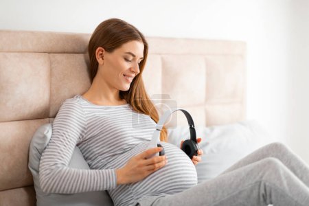 Smiling pregnant lady gently cradling her belly with one hand and holding headphones with the other, expressing tenderness
