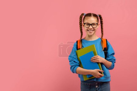 Ein fröhliches Schulmädchen mit Zöpfen, blauen Büchern und einem Rucksack lächelt in die Kamera
