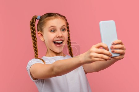 An excited young girl takes a selfie with her smartphone, looking thrilled on a pink background