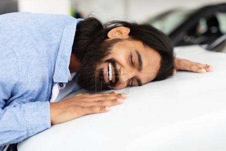 Téléchargez les photos : Un Indien extatique dans une chemise en jean embrasse une nouvelle voiture blanche avec affection dans un showroom de voiture lumineuse, transmettant sa joie et son attachement au nouveau véhicule - en image libre de droit