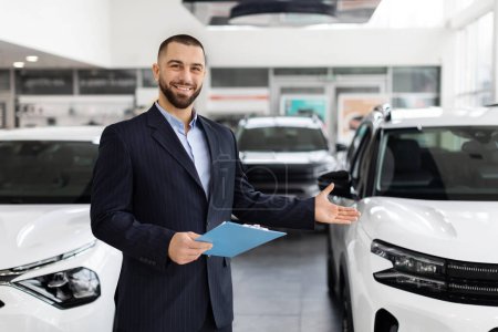 A professional car salesman bearded man in a suit welcomes customers with open arms in a modern car dealership
