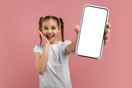 Téléchargez les photos : Une jeune fille joyeuse présente un écran de smartphone vierge prêt pour la personnalisation sur un fond rose - en image libre de droit