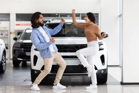 Foto de Una alegre pareja india celebra posiblemente la compra de un coche nuevo en una sala de exposición con un SUV blanco en el fondo - Imagen libre de derechos