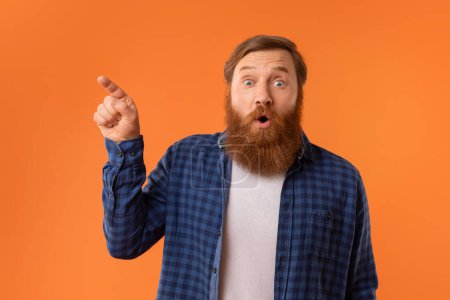Téléchargez les photos : Drôle de portrait studio de l'homme roux surpris avec barbe en tenue décontractée, pointant de côté avec un regard étonné, montrant l'espace de copie sur fond orange. Regardez là concept - en image libre de droit