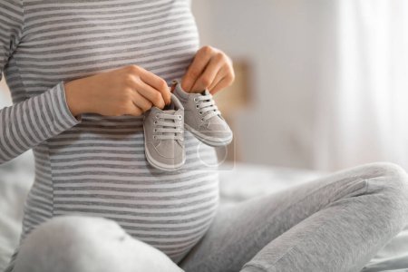 Téléchargez les photos : Une future mère berce une paire de chaussures de bébé près de son ventre, symbolisant la préparation pour la nouvelle arrivée - en image libre de droit