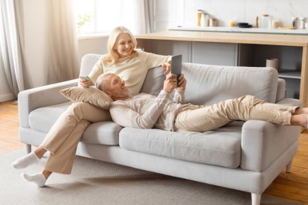 Photo for Mature man holds a tablet while woman extends her leg on a sofa, expressing a relaxed lifestyle - Royalty Free Image