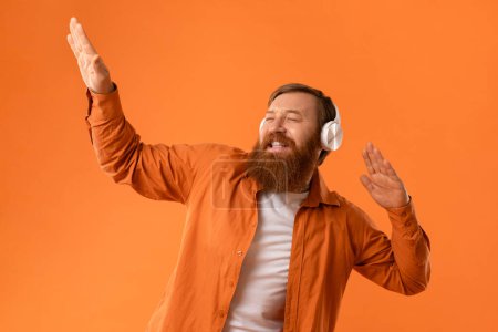 Foto de Retrato del hombre barbudo pelirrojo despreocupado que usa auriculares inalámbricos mientras escucha música y baila levantando brazos sobre el fondo del estudio naranja, cerrando los ojos y cantando con alegría - Imagen libre de derechos