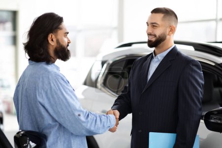A professional salesperson in a suit and a customer indian man shaking hands, sealing a deal in a well-lit modern car dealership showroom