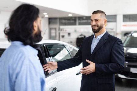 Ein professioneller Autoverkäufer im Anzug unterhält sich in einem Autohaus mit einem potenziellen Kunden.