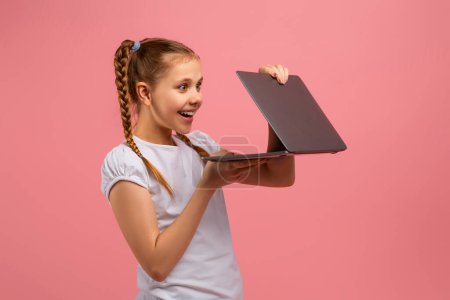 Foto de Una joven sosteniendo un portátil con una expresión sorprendida y encantada sobre un fondo rosa - Imagen libre de derechos