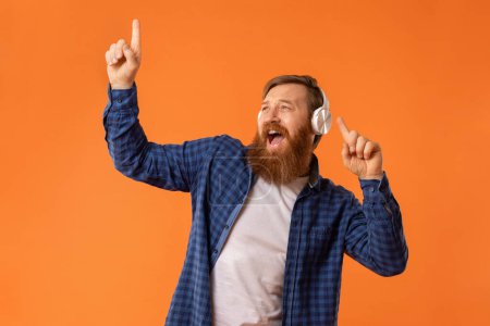 Photo for Portrait of funny guy with read hair and beard singing while listening to music in wireless headphones, over orange studio wall. Joyful man chilling and relaxing, sings his favorite songs - Royalty Free Image