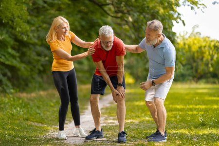 Besorgtes Senioren-Paar hilft seinem männlichen Freund bei der Genesung von Verletzungen beim Outdoor-Training, älterer Mann erleidet Muskelkrämpfe oder Knieverletzungen nach dem Joggen im grünen Park, freier Platz